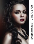 Small photo of Gothic portrait of young green-eyed woman with black curles, black necklace black wings and brown smoky eyes and bardic lipstick standing half turned to the right and looking at you black background