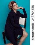 Small photo of Two-third length fashion portrait of girl with lilac smoky eyes and natural lipstick, soft curls, bardic headband and severe dark coat seating half-turned on white leather chair on blue background