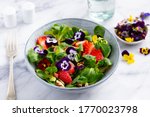 Fresh green salad with strawberries and edible flowers in a bowl. Marble background. Close up.