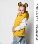 Small photo of Studio shot of little girl in warm outwear yellow sleeveless vest, jeans and sweatshirt turning with her face and looking at camera isolated over white background. Children trendy outfit and fashion