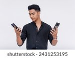 A young Asian man appears indecisive while comparing two smartphones, isolated on a clean white background. Cellphone shopping and specification comparison concepts.
