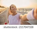 Small photo of Sportswoman, female athlete in pink t-shirt and wristbands smiles holding a mobile phone in her outstretched hands while making self-portrait during morning jog and workout on the bridge at sunrise