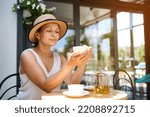 Attractive Latin American woman of natural beauty, wearing a straw hat, sitting at a table in the summer terrace, dreamily looking at a white cup of coffee in her hands, enjoying her weekend outdoor.