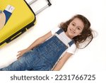Charming European little child, adorable baby girl passenger wearing blue denim overalls, lies on white background near her boarding pass on yellow suitcase, smiles a beautiful toothy smile to camera