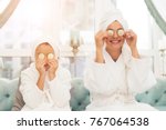 Photo of mother and daughter in white bathrobes. They are sitting on the couch and applying pieces of cucumber to their eyes. Their hair is wrapped in a white bath towel.