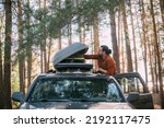 A young man opens the upper tourist trunk of a car in a pine forest on the shore of a lake at sunset. A tourist and an SUV with a trunk on the roof among the trees on a forest road in the sunlight.