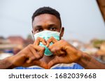 Small photo of young handsome african nigerian man wore face mask preventing, prevent, prevented himself from the outbreak in his society and did a love sign.
