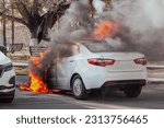 Small photo of Burning car. Fire of a white passenger car in a city parking lot. Fire in the engine compartment, short circuit in the wiring. Open fire and black smoke. Road incident
