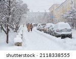 Snow covered city street during a heavy snowfall. Lots of snow on the sidewalk, cars and tree branches. Women walk around the winter city. Cold snowy weather. Magadan, Siberia, Russian Far East.