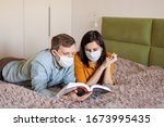 A young couple is sitting at home on self-quarantine. Husband and wife quarantined  coronavirus in protective masks. New reality. Normal life in isolation. Read a book together. Italy europe covid-19