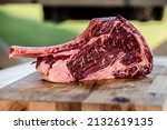 Small photo of Dryaged raw Tomahawk steak on wooden background with vegetables for grilling. Premium beef grilling on a kamado grill. Bone-in ribeye steak on wooden cutting board grilling on coals. Premium steak cut