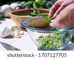Close up of the hands of a woman shelling peas from the pod inside a glass bowl outdoors with sunlight