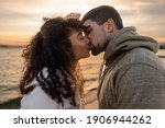 Small photo of Head and shoulders portrait of young beautiful couple in love kissing at sunset in winter seaside resort with cloudy sky. Two millennials in vacation travel manifesting their heart sentiments outdoor
