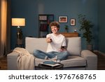 A teenager, a brunette with curly hair relaxes on the living room couch in the evening. The young boy holds the remote control in his hand and switches the channel to a funny program