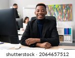 Small photo of Sympathetic smiling new corporate office worker, male wearing carny jacket and turtleneck, looks into camera with big brown eyes, white teeth, dark skin, working behind desk in front of computer