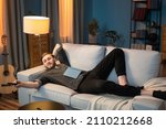 Small photo of Adult man is resting after work in the evening in living room, lying along the couch, propped up by head, leg bent at the knee, a book lying on stomach, switching channels with the TV remote control
