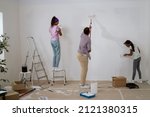 Small photo of A young father and daughters renovate an apartment in the absence of his wife, surprise, they hold rollers with white paint and precisely painting the walls, a girl climbs a ladder