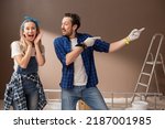Small photo of Couple in love are renovating house after moving in, painting walls in room, decorating the apartment, man is smiling and pointing fingers at empty place, wife is looking at camera excitedly.