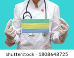 Small photo of Epidemic in Gabon. Young woman doctor in a medical coat or suit and gloves holds a medical mask with the print of the flag of Gabonese Republic on a blue background isolated