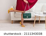 A beautiful young woman poses in a red dress against the background of a sofa in the living room. A woman is holding a green leather shoulder bag. The concept of women's fashion.