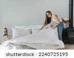 Laughing caucasian beautiful young woman makes bed wears jeans and white t-shirt and glasses. Pretty hispanic girl makes housekeeping. Mockup, people at home. Cheerful student makes order home.