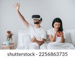 Small photo of Serious young adult woman sitting on bed holds phone watching news while her boyfriend playing games using virtual reality headset. Couple in bed at morning. Bored woman using smartphone. Disunity