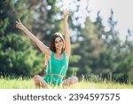 Young cheerful brunette girl in green sportswear sitting in lotus pose in green grass outdoors looks at camera smiles riding hands happily. Fit caucasian young woman trains at park, sunny day