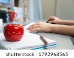 A selective focus shot of a kitchen timer in the form of a tomato on a notebook with a female working on her computer in the background