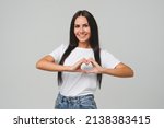 Romantic caucasian young woman in white T-shirt showing heart gesture for love and care relationship, charity, cardiovascular diseases treatment isolated in grey background