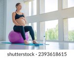 Happy pregnant woman sitting on a fitness ball and touching her belly with look in window. Caucasian expectant mother doing supportive physical exercises for smooth pregnancy