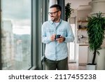 Small photo of Smiling young adult businessman holding drinking coffee cup in office entrepreneur looking through window while standing in modern workplace man taking a time off or taking a brake from hard work