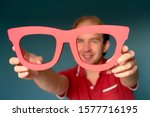 Cheerful man in a red shirt holding big red glasses and looking through . Good Vision Concept. Approach to exaggeration and distortion of facts
