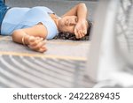 Small photo of A woman lies on the floor, with a fan nearby, seeking respite from the heat with a makeshift cooling solution.