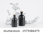 Small photo of Blank cosmetic glass black bottles with beige bungs decorated silver leaves on light white wood table as mock up for advertising, branding identity, design.