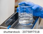 Small photo of Scientist in blue rubber puts a beaker for dissolving or cleaning into an ultrasound bath. Clinical, toxicological and forensic analysis.