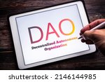 Small photo of The sign of DAO (Decentralized Autonomous Organization), an organization represented by rules encoded as a transparent computer program, on tablet on wooden table. Man hand holding wireless stylus