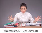 Waist-up portrait of girl sitting at the table with documents and an alarm clock, breeding her hands because she has no time, confused looking at you, isolated on grey background with copy place