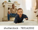 Happy childhood. Happy dark-haired afro-american man laughing and watching his cheerful young son crawling on the floor