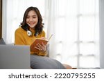 Beautiful young Asian female in casual clothes writing something on her notebook while looking at laptop screen, sitting in her bedroom or living room. online learning, work from home, remote working