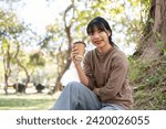 A relaxed and happy Asian woman in casual clothes is sipping coffee while sitting under a tree in a park, enjoying her free time alone. people and lifestyle concepts