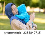 Small photo of Cute young islamic kazakh woman hugging her little crying son, newborn boy. Yappy asian mother in hijab soothes, holding baby in hands outdoors. Family concept