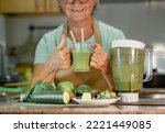 Mature senior woman drinking healthy green smoothie holding a glass with straws. Smiling caucasian lady in home kitchen drinking detox juice