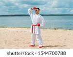 Small photo of Karate man in a sports kimono and red gloves with a red belt in training. Raising his hand to his forehead, he squints in the sun. The action takes place against the backdrop of sand and sea