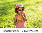 Small photo of Portrait of little Black girl drinking orange juice in a glass with straw at outdoor park. Child in summer hat suck from the straw a fresh juice