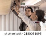 Businessman traveller putting luggage in overhead compartments with female flight attendant helping, cabin crew woman working on board, female airhostess helping passenger put suitcase in compartment