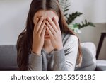 Small photo of Lost in thoughts stressed brunette female regretting of wrong decision. Close up of unhappy anxiety young caucasian female covering face with hands sitting on grey couch in living room at home