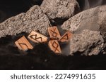Small photo of the wooden rune of Inguz among other runes, symbols of the earth, lie in the ground amid smoke and large stones. Mysticism and the rite of fruitful earth