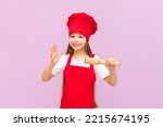 A little girl in a chef's hat is preparing for baking, holding a rolling pin in her hands to roll out the dough. Baby baker on a pink isolated background.