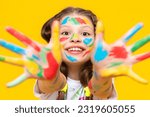 Small photo of Childish pranks. A charming child with painted palms with multicolored paints. A little girl happily draws with her hands. Yellow isolated background.