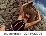 a portrait of a woman in a swimsuit, hat and sunglasses in summer on the riverbank by a concrete wall